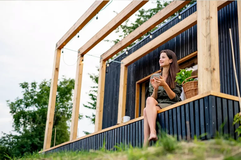 Frau sitzt mit Tasse auf der Veranda eines modernen Tiny Houses im Grünen