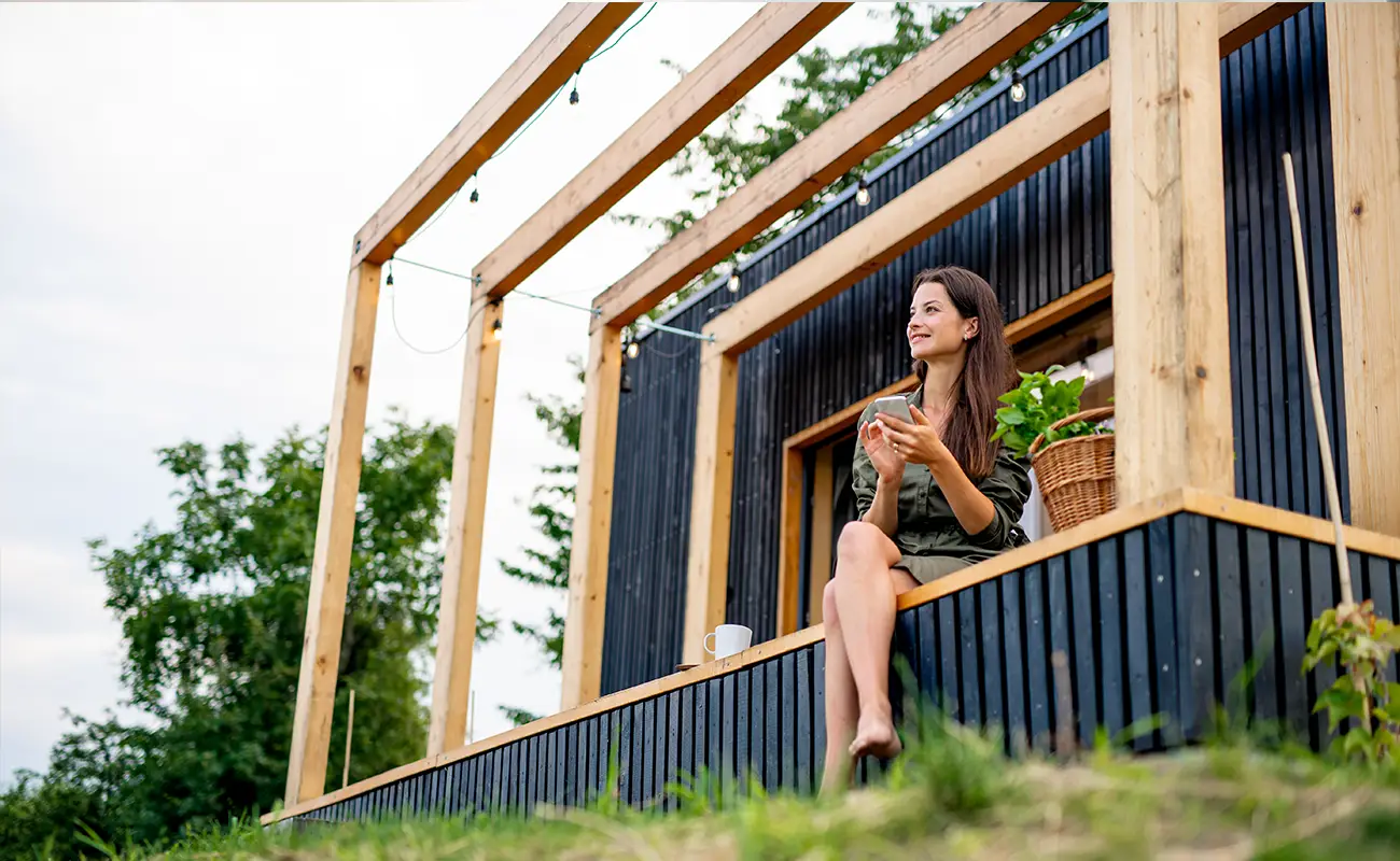 Frau sitzt mit Tasse auf der Veranda eines modernen Tiny Houses im Grünen