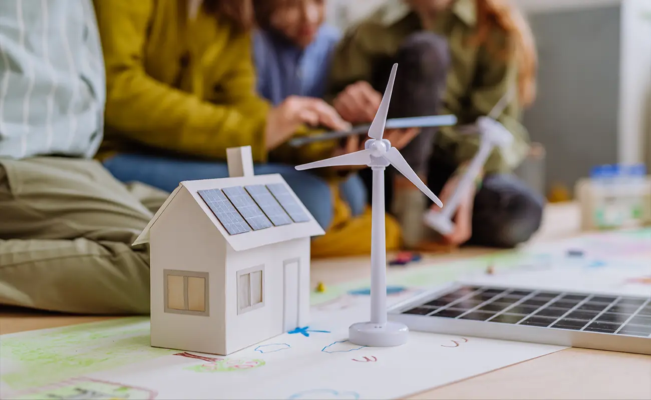 Miniaturhaus mit Solardach neben Windrad, Kinder spielen im Hintergrund mit Lernmaterial.