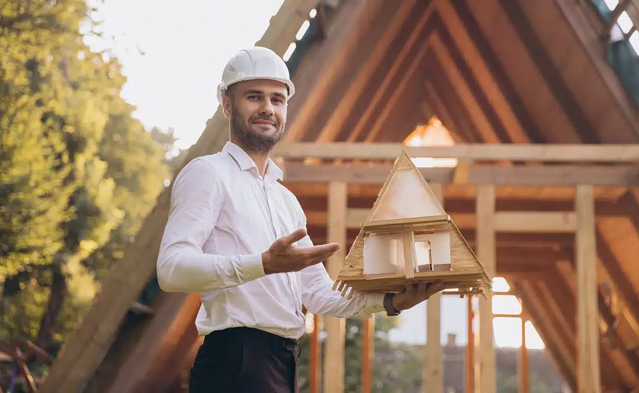 Architekt mit Schutzhelm hält Modell eines Holzhauses vor Rohbau in Dreiecksform.