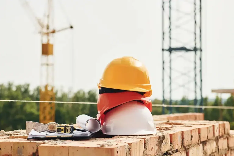 Schutzhelme, Baupläne und Maßband auf Ziegelmauer mit Baukränen im Hintergrund.